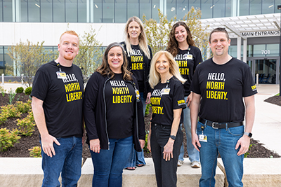 Group of University of Iowa hospital staff smiling outside the main entrance, wearing matching black custom t-shirts with bold yellow text reading “Hello, North Liberty.” Bulk order apparel created for hospital staff unity and community outreach.